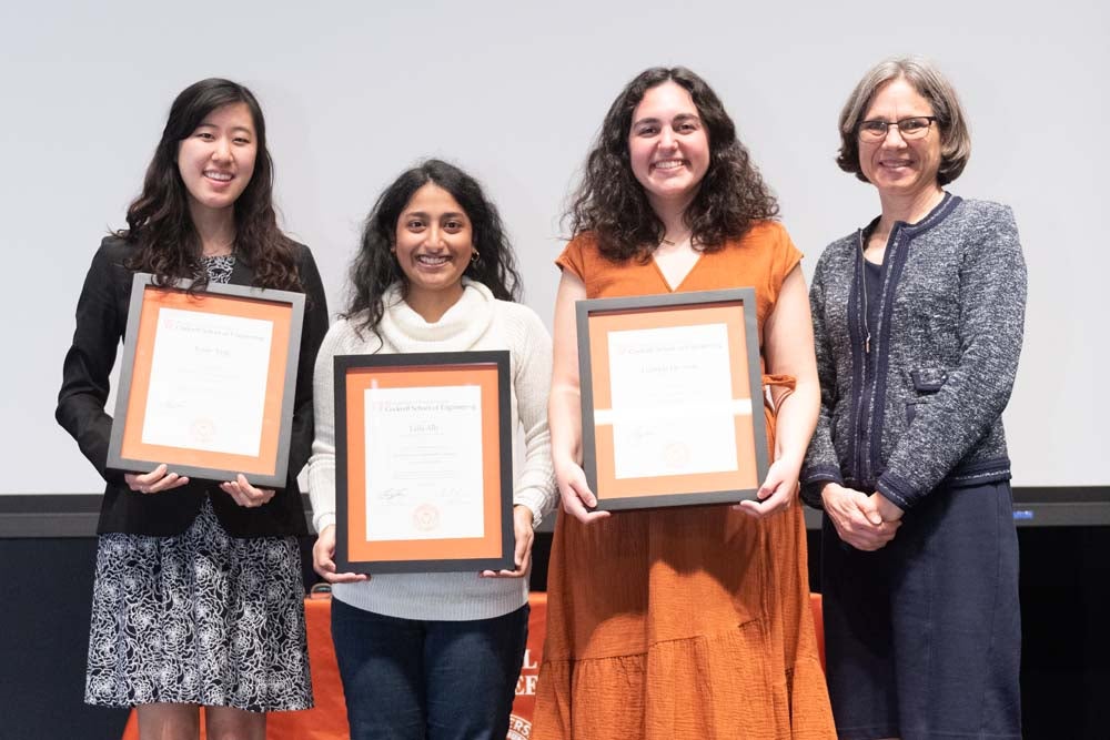 three students standing on stage holding frames next to Dean Sharon Wood
